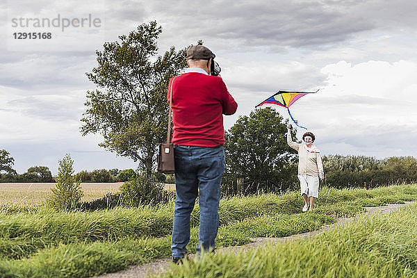 Senior man filming wife flying kite in rural landscape