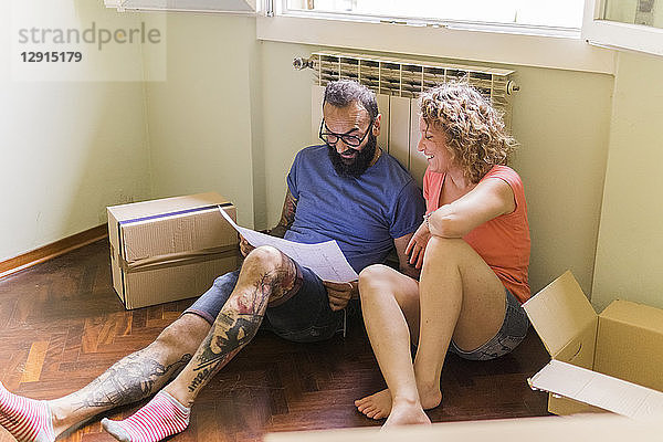 Happy couple sitting side by side on the floor of new home looking at plan