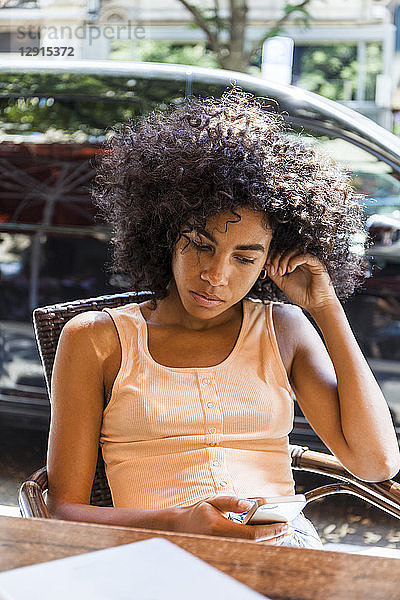 Portrait of young woman with curly hair sitting at sidewalk cafe looking at cell phone