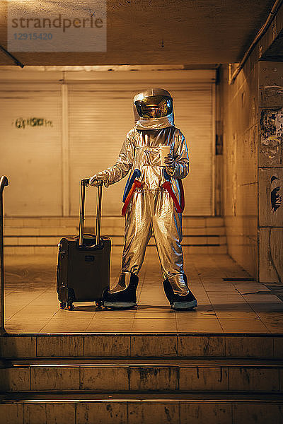 Spaceman in the city at night standing in underpass with rolling suitcase and takeaway coffee
