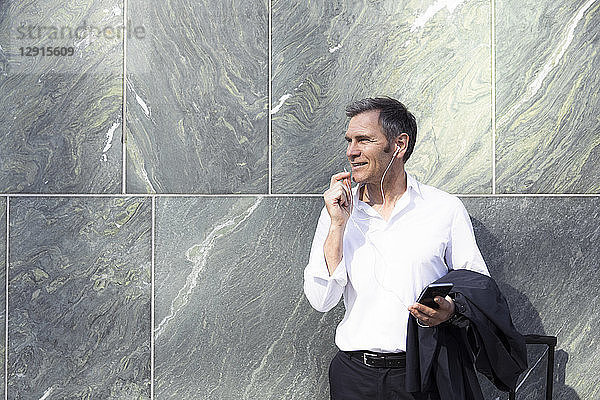 Mature businessman with earphones and smartphone at a wall
