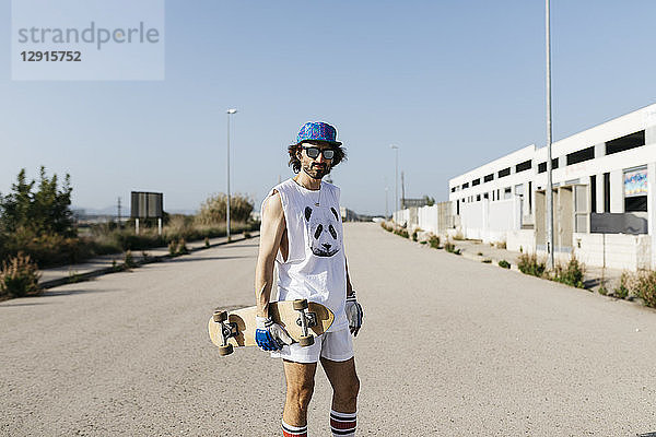 Portrait of sportive man in white and blue with skateboard on empty gray paved road