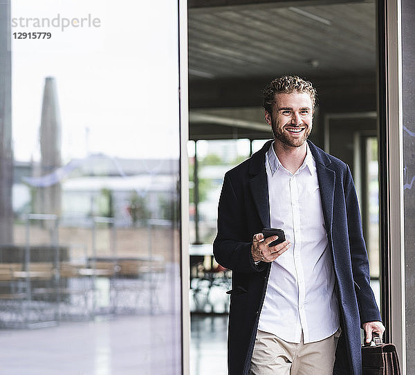 Smiling young businessman holding cell phone leaving office