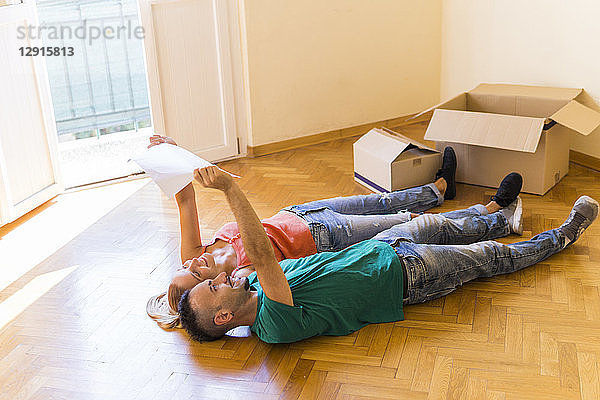 Happy couple lying side by side on the floor of new home looking together at contract