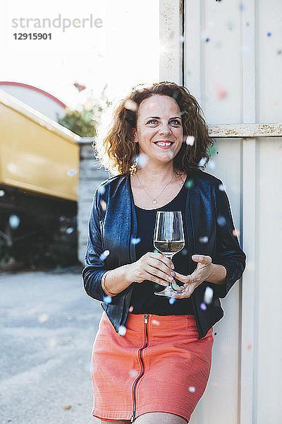 Portrait of smiling woman with glass of white wine looking at flying confetti