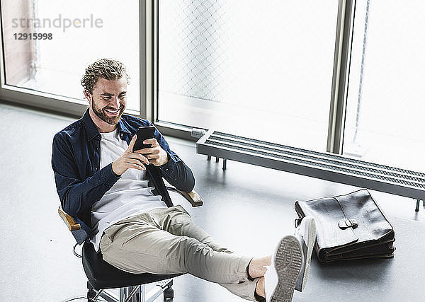Smiling casual businessman sitting in office with feet up using cell phone