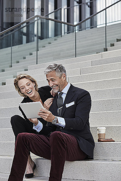 Portrait of two businesspeople sitting together on stairs looking at tablet having fun