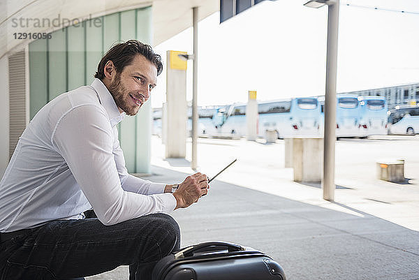 Smiling businessman with suitcase sitting at bus terminal using tablet