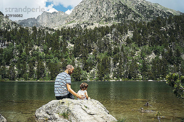 Spain  Father and daughter sitting on a rock at a mountain lake  feeding ducks