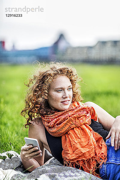 Portrait of smiling young woman with curly red hair wearing orange scarf lying on blanket on a meadow