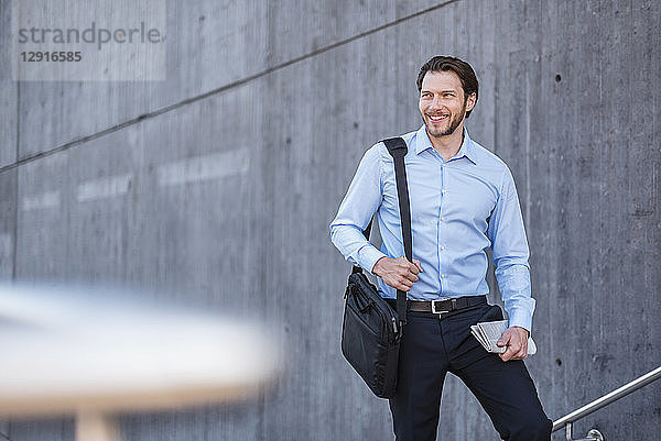 Portrait of smiling businessman with laptop bag at concrete wall
