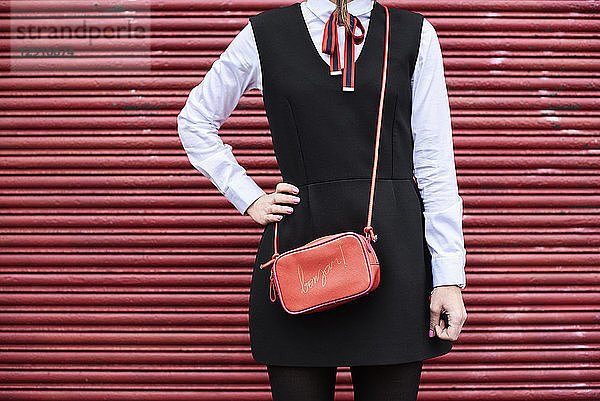 Fashionable woman with red handbag wearing black dress standing in front of red roller shutter