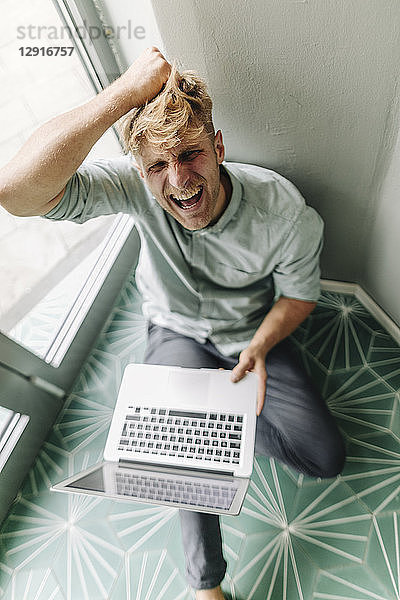 Young man sitting on ground  holding laptop  screaming in despair
