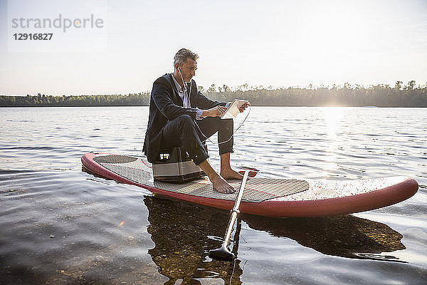 Businessman sitting on paddleboard on a lake using tablet and earphones