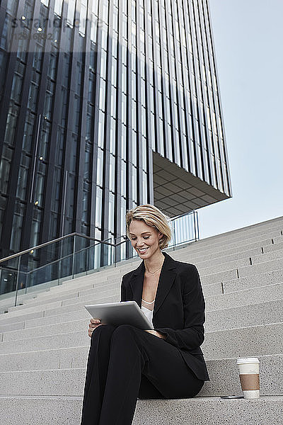 Blond businesswoman with coffee to go sitting on stairs in front of modern office building using tablet