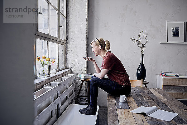Woman sitting on desk in loft using cell phone
