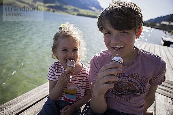 Austria  Tyrol  Walchsee  portrait of brother and sister sitting on jetty at a lake eating ice cream cones
