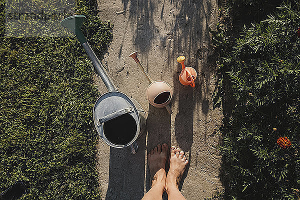 Three watering cans and woman's feet standing on garden path  top view