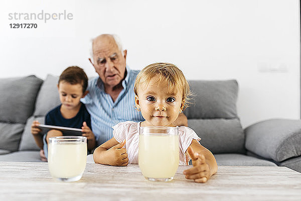Portrait of little girl with glass of lemonade at home with brother and grandfather in the background