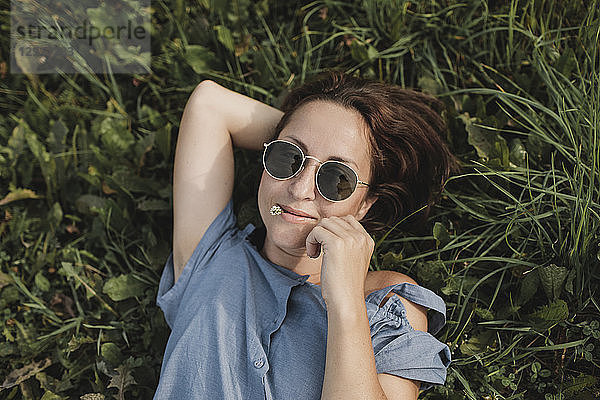 Portrait of smiling woman wearing sunglasses lying in grass with flower in her mouth