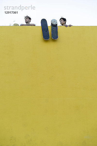Two young men with skateboards behind a high yellow wall looking around