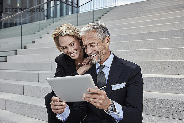 Portrait of two laughing businesspeople sitting together on stairs looking at tablet