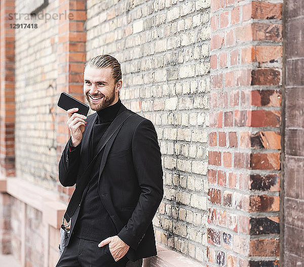 Smiling fashionable young man standing at brick wall using cell phone