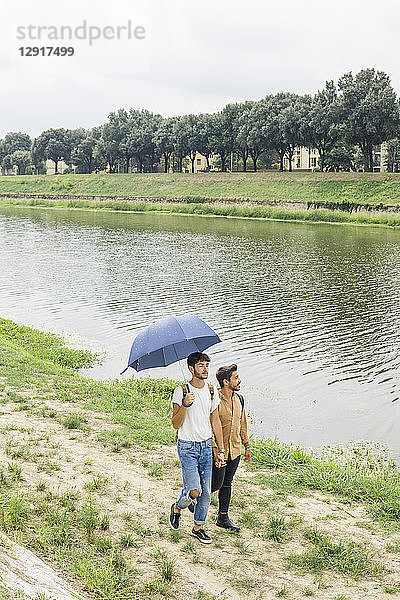 Young gay couple with umbrella strolling hand in hand at riverside