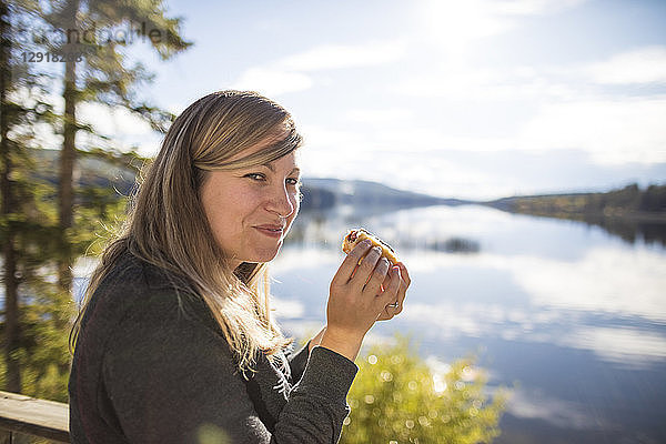Seitenansicht einer lächelnden Frau  die in die Kamera schaut  während sie am Seeufer einen Hotdog isst