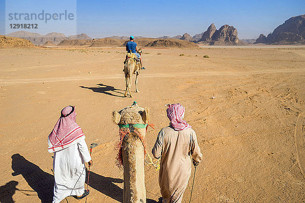 Ich-Perspektive beim Reiten auf einem Kamel durch die Wüste von Wadi Rum  geschützte Wüstenwildnis im südlichen Jordanien  mit Sandsteinbergen und einem Mann auf einem Kamel in der Ferne  Dorf Wadi Rum  Verwaltungsbezirk Aqaba  Jordanien