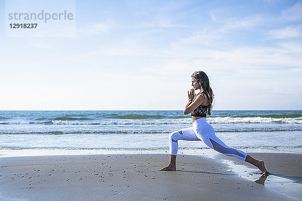Seitenansicht einer jungen Frau mit schwarzen Haaren beim Yoga am Strand