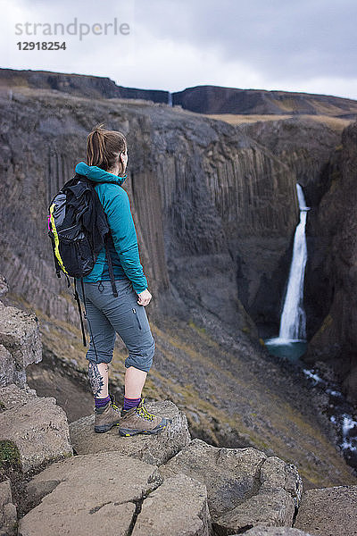 Weibliche Rucksacktouristin steht auf einem Felsen und blickt auf den Litlanesfoss-Wasserfall in der Ferne  Island