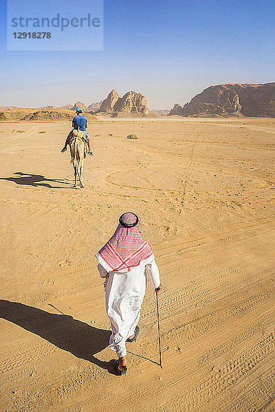 Blick auf einen Mann in arabischer Kleidung  der durch die Wüste von Wadi Rum läuft  geschützte Wüstenwildnis im Süden Jordaniens  mit Sandsteinbergen und einem Mann  der auf einem Kamel reitet  in der Ferne  Dorf Wadi Rum  Verwaltungsbezirk Aqaba  Jordanien