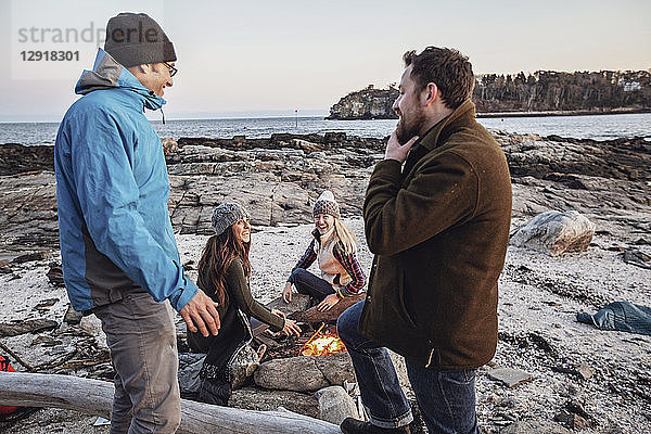 Vier erwachsene Freunde  die sich am Lagerfeuer unterhalten und lächeln  während sie an einem felsigen Küstenstrand im Herbst campen  Portland  Maine  USA