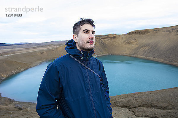 Ein junger Mann in einer blauen Jacke blickt in die Kamera. Im Hintergrund ist der Viti-Explosionskrater im Vulkangebiet Krafla in der Nähe des Myvatn-Sees in Nordisland zu sehen.