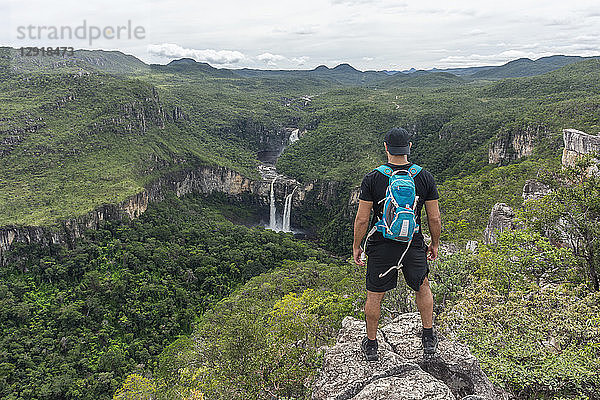 Junger Erwachsener steht auf einem Felsvorsprung mit schöner Cerrado-Naturlandschaft im Hintergrund  Mirante da Janela-Wanderung  Chapada dos Veadeiros  Goias  Zentralbrasilien