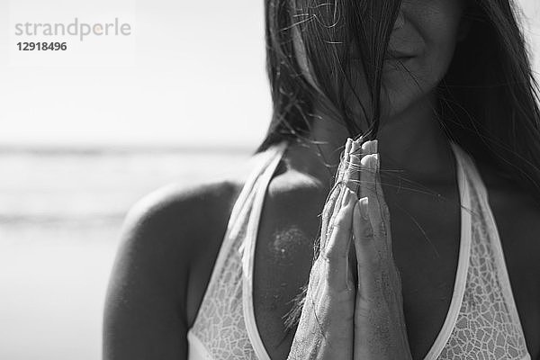 Vorderansicht einer jungen Frau mit schwarzen Haaren und verschränkten Händen beim Yoga am Strand