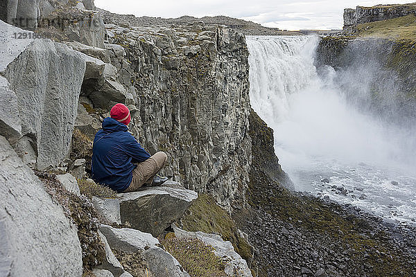 Ein junger Mann mit blauer Jacke und roter Mütze setzt sich auf einen Felsen an der Steilwand und bewundert den Dettifoss-Wasserfall in Nordisland.