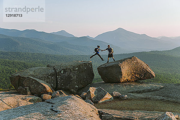 Entfernte Ansicht eines Mannes  der einer Frau beim Sprung über einen Felsbrocken hilft  Pitchoffâ€ Mountain  Adirondack Mountains  New York State  USA