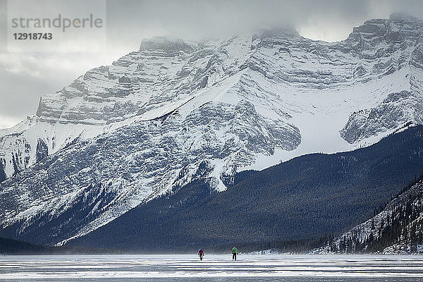 Fernblick auf zwei Personen beim Schlittschuhlaufen auf dem zugefrorenen Lake Minnewanka im Winter  Banff National Park  Alberta  Kanada