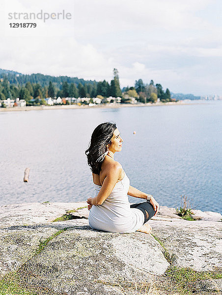 Frau mit schwarzen Haaren sitzt und dreht den Oberkörper während sie Yoga am Meer macht