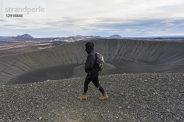 Eine junge  schwarz gekleidete Frau in Stiefeln geht auf dem Kamm des Hverfjall-Kraters entlang  während im vulkanischen Hintergrund Dampf aufsteigt.