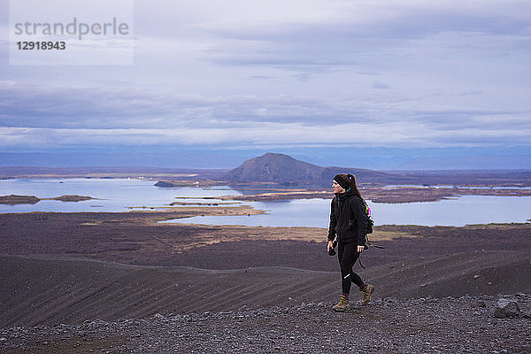 Eine junge  schwarz gekleidete Frau in Stiefeln spaziert auf dem Kamm des Kraters Hverfjall entlang  mit dem Myvatn-See im Hintergrund.