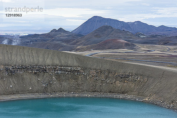 Zwei Personen wandern entlang des Kammes des Viti-Kraters  mit dem blauen See im Vordergrund und dem geothermischen Kraftwerk Krafla  das im Hintergrund Dampf abgibt  in Nordisland in der Nähe des Myvatn-Sees.