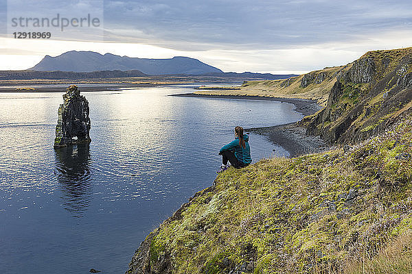 Wanderin sitzt am Rand einer Küstenklippe und betrachtet eine Felsformation  Hvitserkur  Island