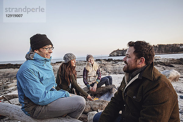 Vier erwachsene Freunde  die sich am Lagerfeuer unterhalten und lächeln  während sie an einem felsigen Küstenstrand im Herbst campen  Portland  Maine  USA