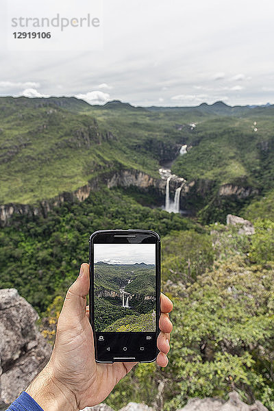 Eine Hand hält ein Handy und fotografiert eine wunderschöne Cerrado-Naturlandschaft mit Wasserfällen in Mirante da Janela  Chapada dos Veadeiros  Goias  Zentralbrasilien