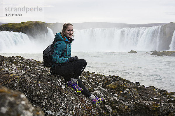 Porträt einer Wanderin  die vor dem plätschernden Godafoss-Wasserfall sitzt und in die Kamera lächelt  Island