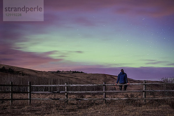 Rückansicht einer auf einem Zaun sitzenden Frau unter Polarlicht am nächtlichen Himmel  Morley  Alberta  Kanada