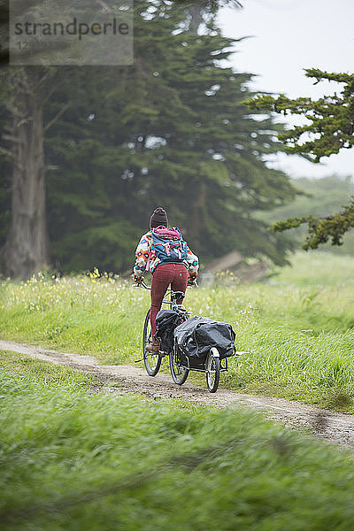 Junge erwachsene Frau fährt mit dem Fahrrad durch eine grasbewachsene Gegend und zieht einen kleinen Anhänger  Santa Cruz  Kalifornien  USA